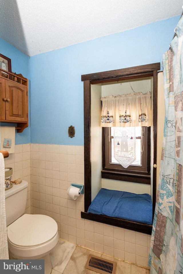 full bath featuring a textured ceiling, toilet, visible vents, tile walls, and wainscoting