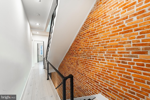 hall with light wood-type flooring, brick wall, baseboards, and an upstairs landing