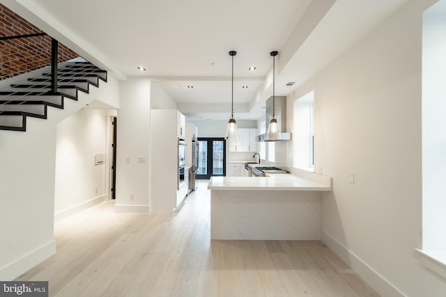 kitchen with light wood finished floors, white cabinets, light countertops, wall chimney range hood, and a sink