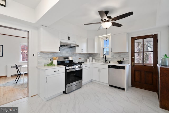 kitchen featuring under cabinet range hood, a sink, light countertops, appliances with stainless steel finishes, and radiator heating unit