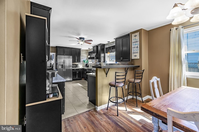 kitchen featuring ceiling fan, a breakfast bar area, a peninsula, dark cabinetry, and stainless steel fridge with ice dispenser