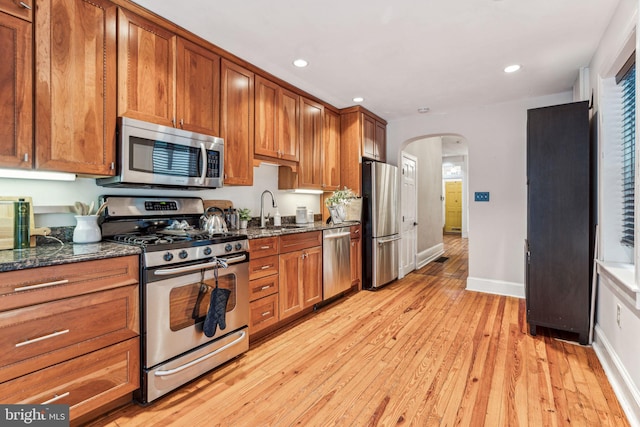 kitchen with arched walkways, stainless steel appliances, light wood-style flooring, brown cabinetry, and a sink