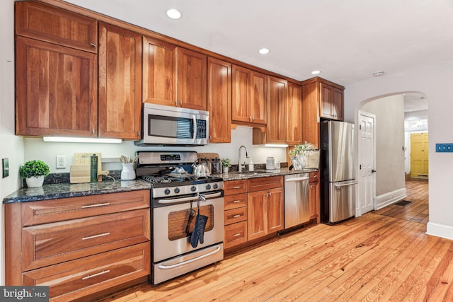 kitchen with arched walkways, light wood-style flooring, a sink, appliances with stainless steel finishes, and dark stone countertops