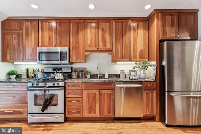 kitchen featuring a sink, appliances with stainless steel finishes, light wood-type flooring, brown cabinets, and dark stone countertops