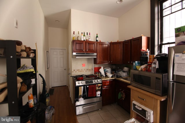 kitchen with light tile patterned floors, under cabinet range hood, stainless steel appliances, backsplash, and reddish brown cabinets