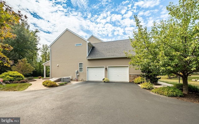 view of side of property featuring driveway, a garage, a shingled roof, stone siding, and central air condition unit