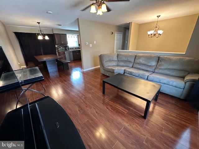 living area featuring ceiling fan with notable chandelier, dark wood-type flooring, and baseboards