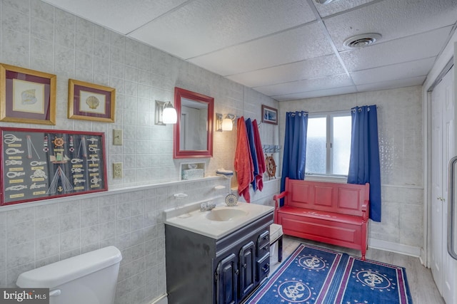 bathroom featuring toilet, vanity, wood finished floors, tile walls, and a paneled ceiling