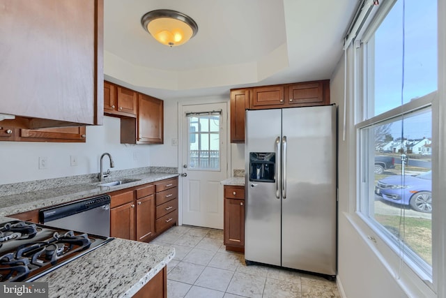 kitchen with light stone countertops, brown cabinetry, a tray ceiling, a sink, and appliances with stainless steel finishes