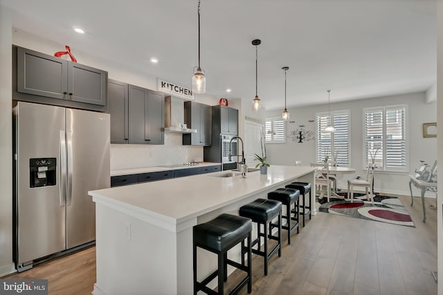 kitchen featuring a kitchen island with sink, a sink, light wood-type flooring, wall chimney exhaust hood, and stainless steel fridge