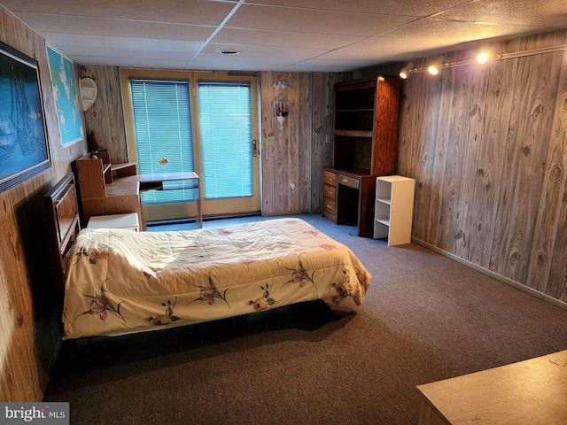 bedroom featuring carpet, a drop ceiling, wooden walls, and baseboards