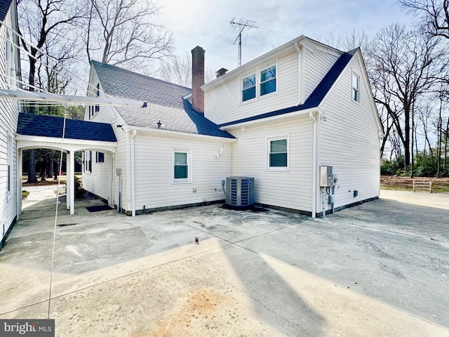 back of house with a shingled roof, a patio area, central AC, and a chimney