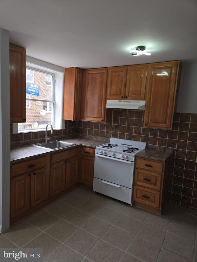 kitchen with brown cabinets, under cabinet range hood, white gas range, dark countertops, and a sink