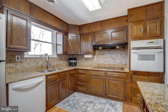 kitchen featuring decorative backsplash, brown cabinetry, a sink, white appliances, and under cabinet range hood