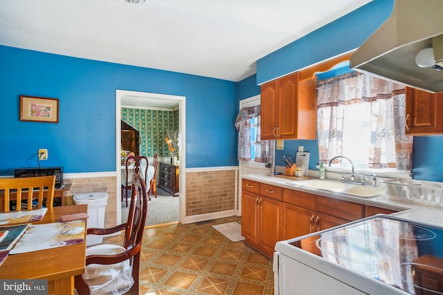 kitchen featuring a sink, under cabinet range hood, white electric stove, brown cabinetry, and wainscoting