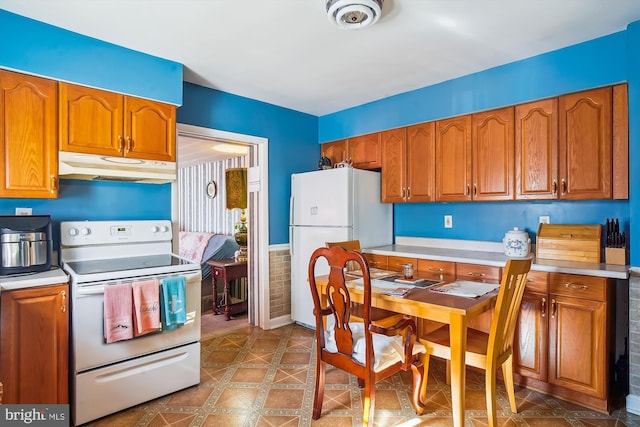 kitchen featuring under cabinet range hood, white appliances, wainscoting, brown cabinetry, and light countertops