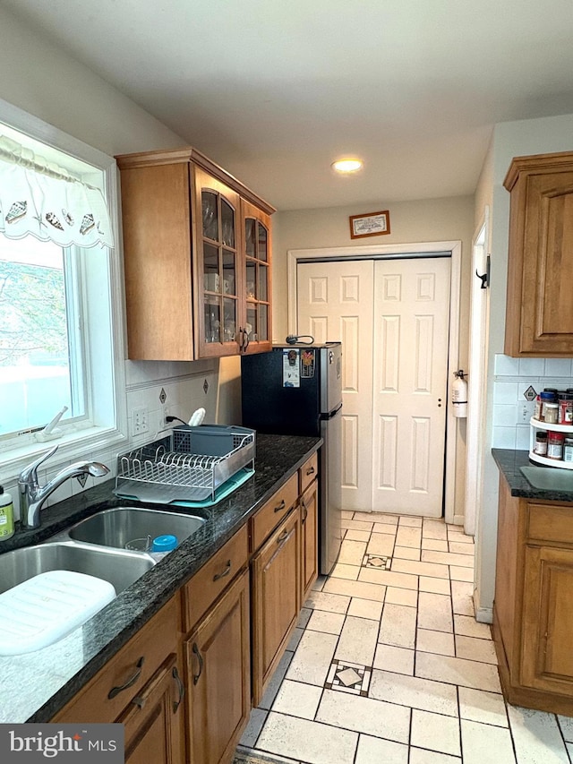 kitchen with a sink, freestanding refrigerator, decorative backsplash, brown cabinetry, and glass insert cabinets