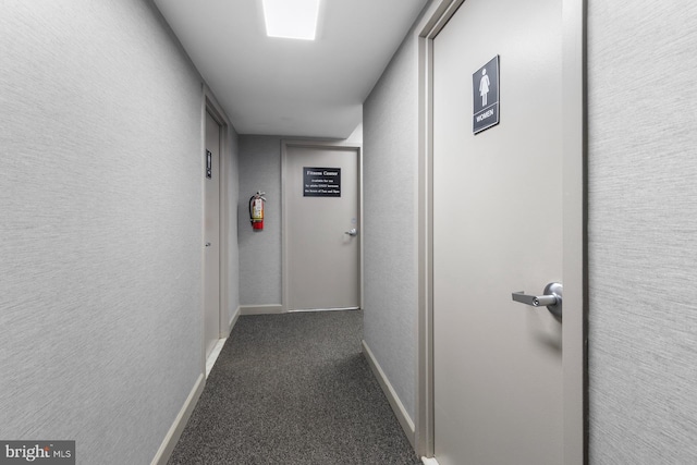 hallway featuring dark colored carpet, a textured wall, and baseboards