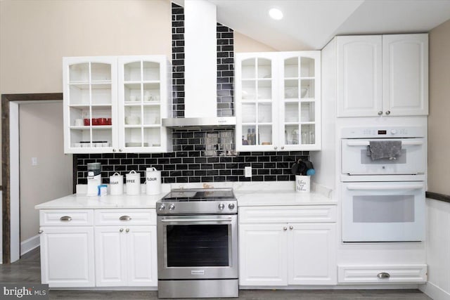 kitchen with stainless steel range with electric stovetop, white double oven, white cabinetry, wall chimney range hood, and backsplash