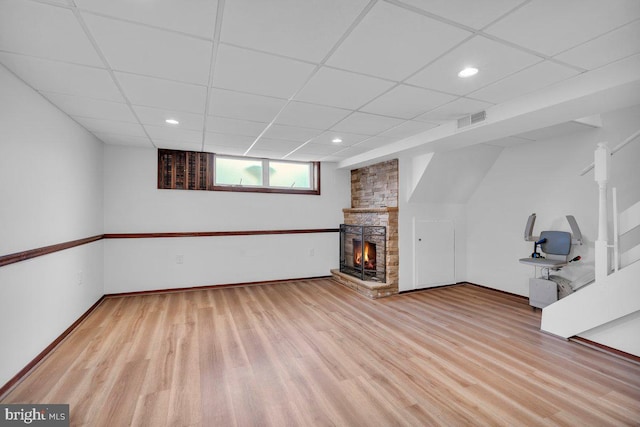 basement with light wood finished floors, a paneled ceiling, visible vents, a stone fireplace, and baseboards