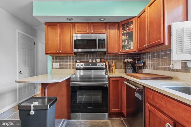 kitchen with brown cabinetry, light countertops, glass insert cabinets, appliances with stainless steel finishes, and tasteful backsplash