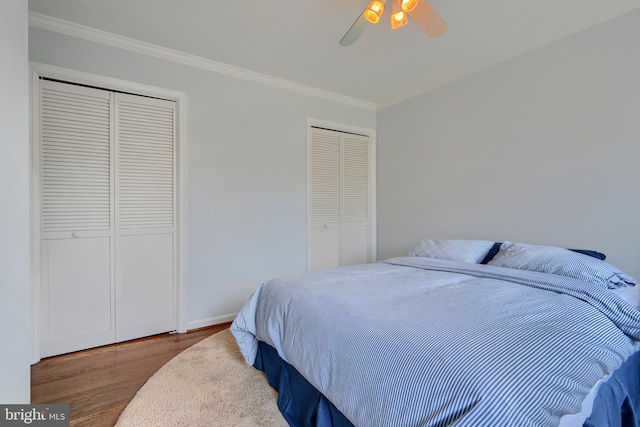 bedroom with two closets, ornamental molding, wood finished floors, baseboards, and ceiling fan