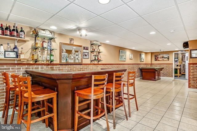 bar with a drop ceiling, wainscoting, brick wall, a bar, and light tile patterned flooring