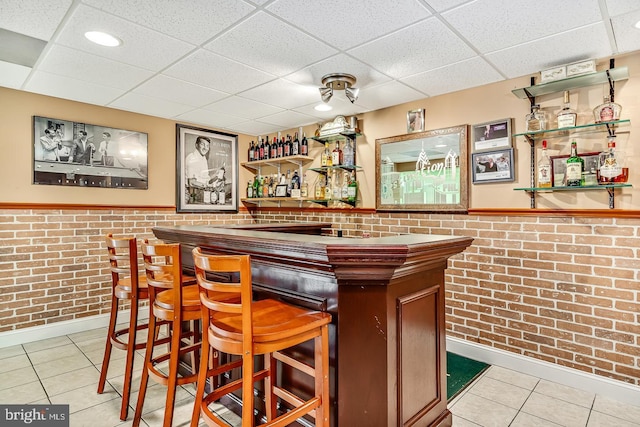 bar featuring a drop ceiling, a wainscoted wall, brick wall, tile patterned floors, and a bar