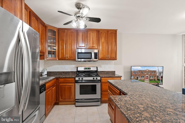 kitchen featuring light tile patterned floors, appliances with stainless steel finishes, brown cabinetry, glass insert cabinets, and dark stone countertops