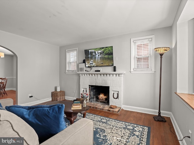 living room with arched walkways, a brick fireplace, baseboards, and wood finished floors