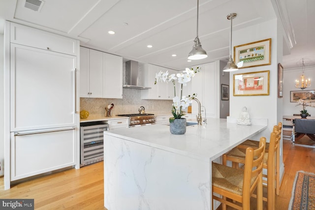 kitchen with a breakfast bar area, beverage cooler, a sink, white cabinetry, and wall chimney exhaust hood