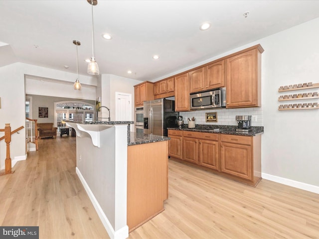 kitchen with light wood finished floors, dark stone counters, decorative light fixtures, stainless steel appliances, and backsplash