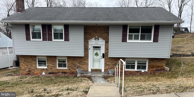 split foyer home featuring roof with shingles, a chimney, fence, and brick siding