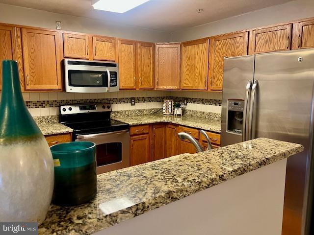 kitchen with brown cabinetry, backsplash, appliances with stainless steel finishes, and light stone countertops