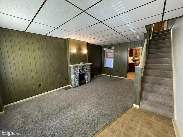 unfurnished living room featuring a stone fireplace, a drop ceiling, wooden walls, carpet floors, and stairway