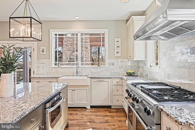 kitchen with light wood finished floors, light stone countertops, stainless steel appliances, wall chimney exhaust hood, and a sink