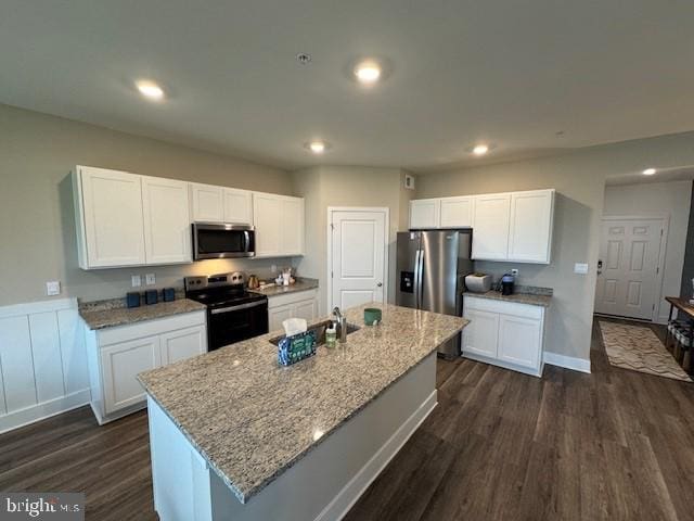 kitchen with dark wood finished floors, a center island with sink, stainless steel appliances, white cabinetry, and light stone countertops