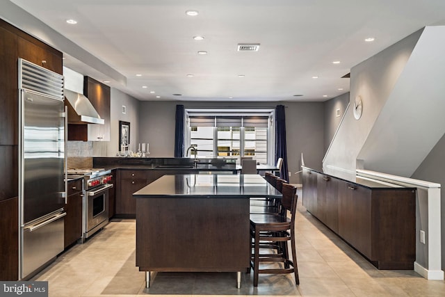 kitchen featuring high end appliances, dark countertops, wall chimney exhaust hood, dark brown cabinets, and a sink