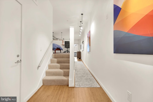 corridor with light wood-type flooring, baseboards, track lighting, and stairs