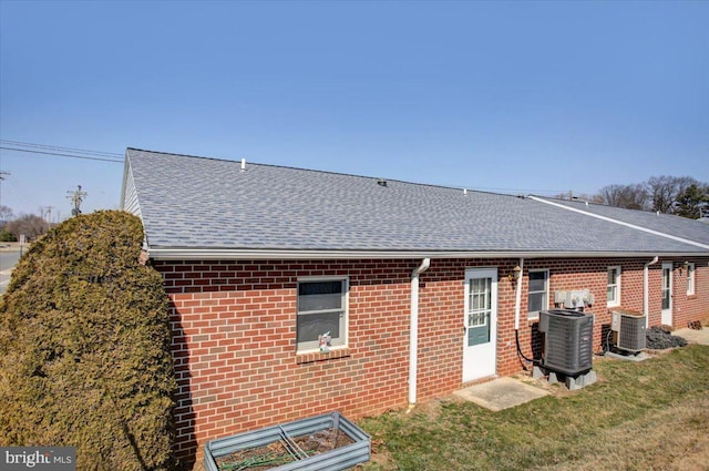 rear view of house with a yard, brick siding, central AC unit, and a shingled roof