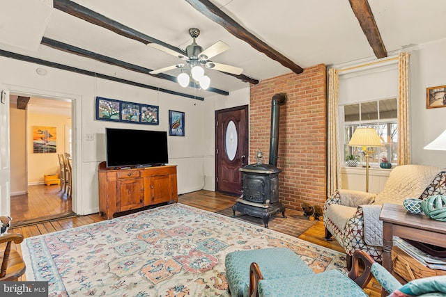 living area with ceiling fan, a wainscoted wall, wood finished floors, beam ceiling, and a wood stove