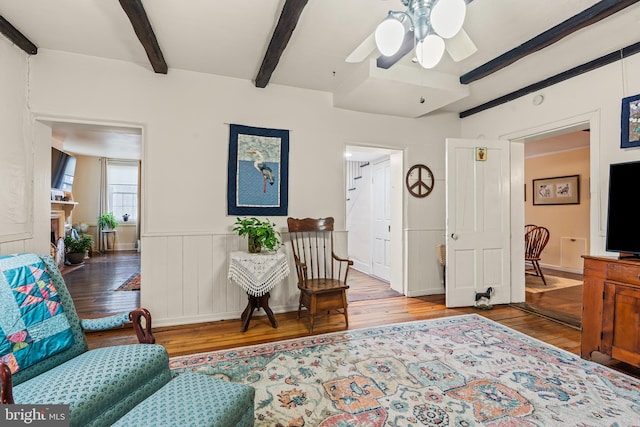 living area with a wainscoted wall, a fireplace, ceiling fan, beamed ceiling, and hardwood / wood-style flooring