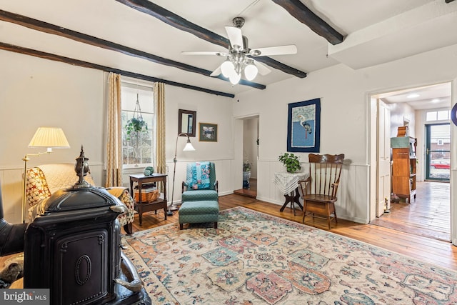 sitting room featuring ceiling fan, wood finished floors, wainscoting, beamed ceiling, and a wood stove