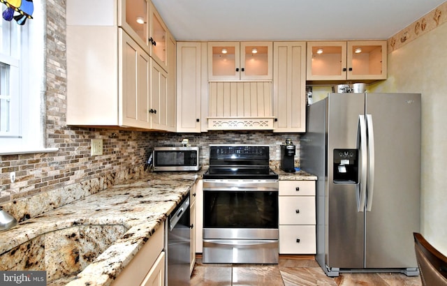 kitchen featuring stainless steel appliances, backsplash, glass insert cabinets, and light stone countertops