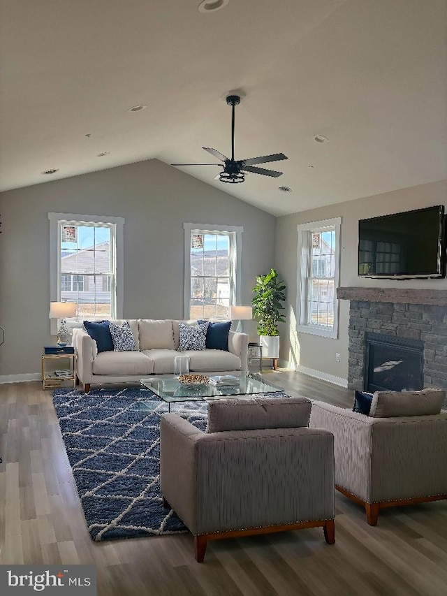 living area featuring ceiling fan, vaulted ceiling, a stone fireplace, wood finished floors, and baseboards