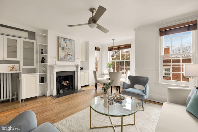 living area with radiator heating unit, ceiling fan, ornamental molding, a fireplace with flush hearth, and light wood-type flooring