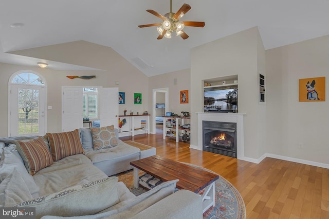 living area with a warm lit fireplace, ceiling fan, lofted ceiling, wood finished floors, and baseboards