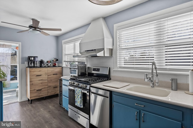 kitchen featuring blue cabinetry, a sink, dark wood finished floors, appliances with stainless steel finishes, and wall chimney range hood