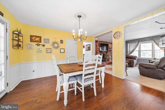 dining area featuring wainscoting, a brick fireplace, an inviting chandelier, and wood finished floors