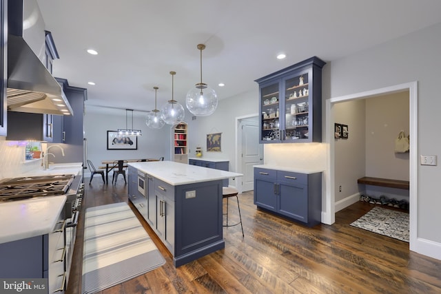 kitchen featuring blue cabinetry, a kitchen island, glass insert cabinets, wall chimney exhaust hood, and dark wood-style flooring